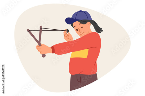 Little mischievous girl shoots with slingshot, closing one eye to aim at target during weekend. Child misbehaves by shooting at peers or glass with homemade slingshot with stretched rubber band.