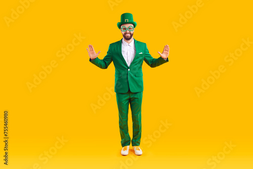 Portrait of a happy man in a leprechaun costume. Full body shot of a funny guy wearing a green suit, hat and shamrock party glasses standing on a yellow studio background. St Patrick's Day concept