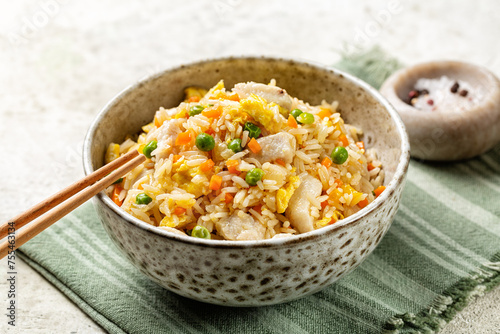 Bowl with fried long rice, chicken, egg and vegetables, onion, bell pepper, carrot, green peas. Beige table with napkin.