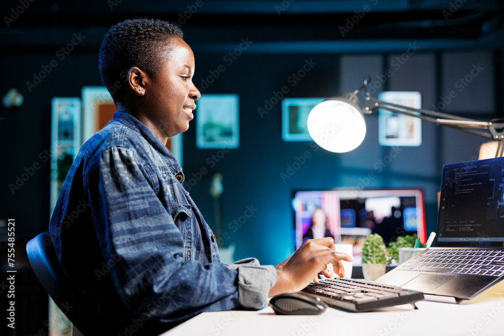 Fototapeta premium Female freelancer works at a desk, using a laptop to manage a futuristic information system. African american woman using her personal computer for cybersecurity and programming, working at home.