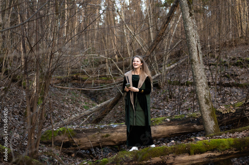 A young woman with a mobile phone stands on a green mossy tree trunk in the forest, early spring