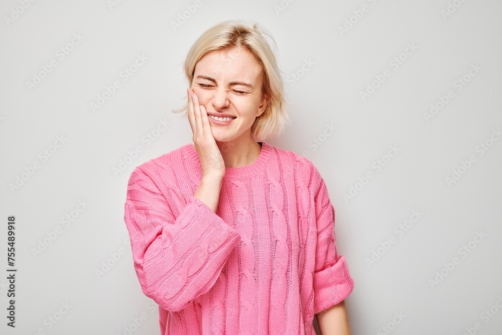 Smiling woman in pink sweater with hand on cheek against a gray background, expressing toothache. Smiling woman in pink sweater with hand on cheek against a gray background, expressing toothache.