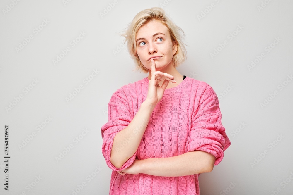 Thoughtful young woman in pink shirt looking up with finger on chin against a gray background. Thoughtful young woman in pink shirt looking up with finger on chin against a gray background.