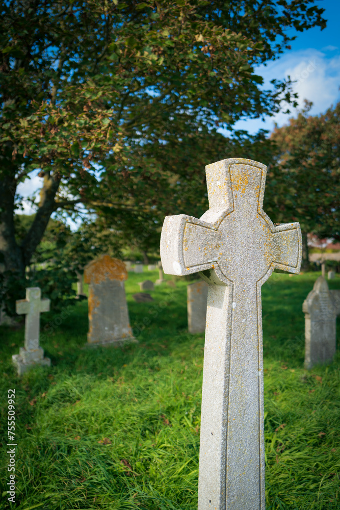 Vertical aspect of an old crucifix shaped headstone seen in a late summer churchyard in England. The lush grass and various headstones can be seen in the background. Vertical aspect of an old crucifix shaped headstone seen in a late summer churchyard in England. The lush grass and various headstones can be seen in the background.