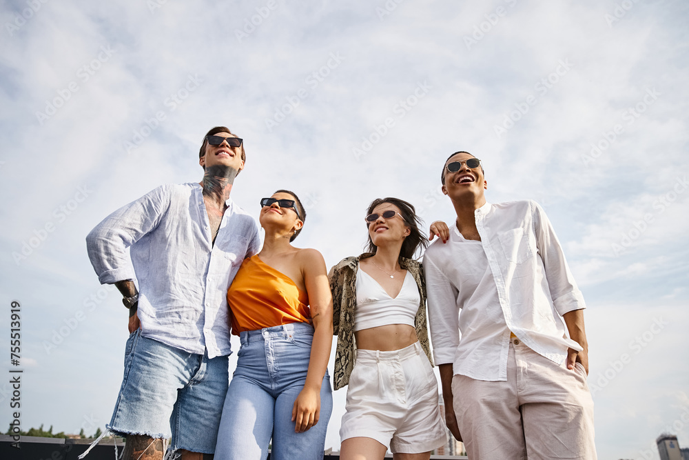 four diverse attractive cheerful friends with stylish sunglasses posing joyfully on rooftop together