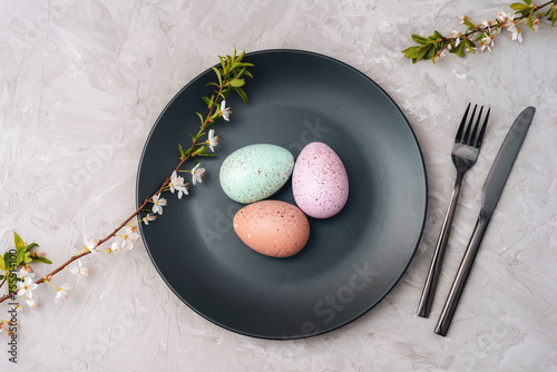 Black plate with Easter eggs, cherry blossom branches and cutlery on textured gray background. Top view, flat lay
