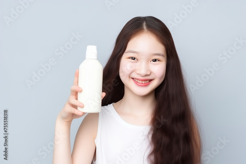 young Asian girl with brown hair and white skin is smiling and holding elegant skincare, whitening bottle in her hand, mockup, on gray