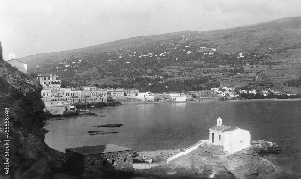 Black and white photo from the island of Andros, Greece, in the 1950s. The edge of the capital town of Andros towards the sea is seen,with small white church in foreground and mountain in the distancw