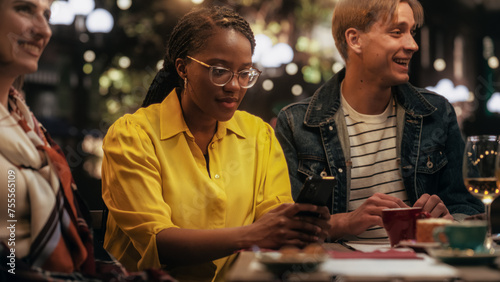 Young Black Female Focused On Smartphone, Not Paying Attention to a Group of Friends Sitting in a Cafe Terrace Around Her. African Girl is Zoned Out, Entertained by Online Social Media on Her Device 