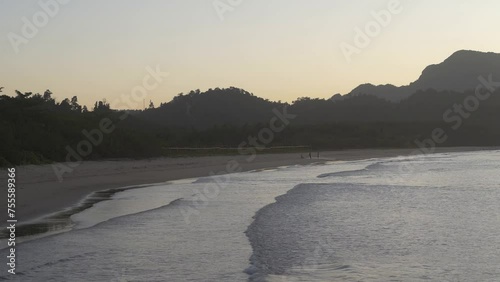 Evening by the ocean. The waves roll towards the beach and spread out on the sand. In the distance, people can be seen folding fishing nets. Mountains against the sky, illuminated in golden.