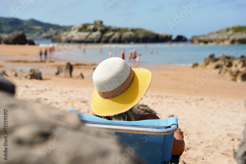 A woman is sitting in a beach chair with a yellow hat sunbathing