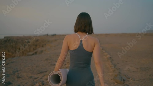 Woman walking with yoga mat on the edge of the cliff by the beach at sunset, Fuerteventura, Spain