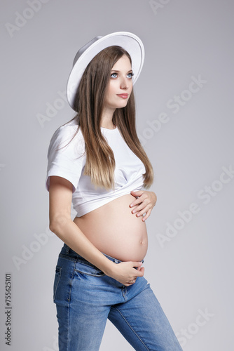 Young pretty pregnant woman in white t-shirt, hat and jeans. Female with hands on belly exposed 5th month of pregnancy