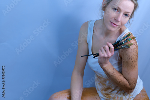 beautiful blonde woman painting in front of a white wall, smiling and holding many brushes in her hand
