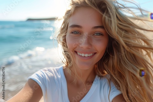 A beautiful girl with blonde hair smiles and takes a selfie while riding on the seashore, wearing a white t-shirt, her long wavy hair fluttering in the wind.
