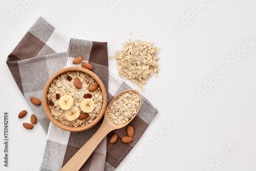 Oatmeal in a wooden bowl with almonds and pieces of banana, scattered oat flakes and with spoon on white background. View from above. Place for text