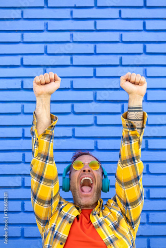 An elated man with headphones and sunglasses shouts in triumph, fists pumped in the air, against a striking blue brick wall