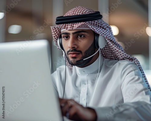 An attentive man dressed in traditional Saudi attire with a headset is working on a computer in a professional setting. photo