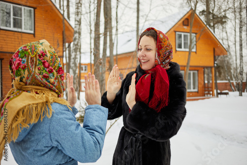 Smiling woman and teenage girl in folk style shawls stand against wooden houses and play okie-dokie on winter day photo