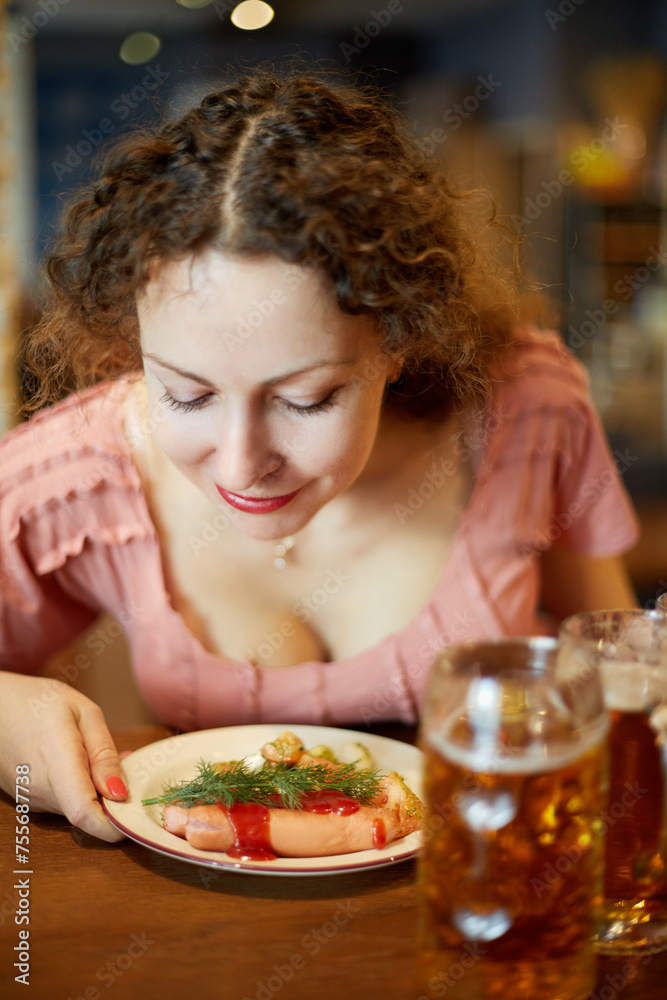 Smiling woman with curly hair sniffs fried sausages with sprig of dill on plate in cafe