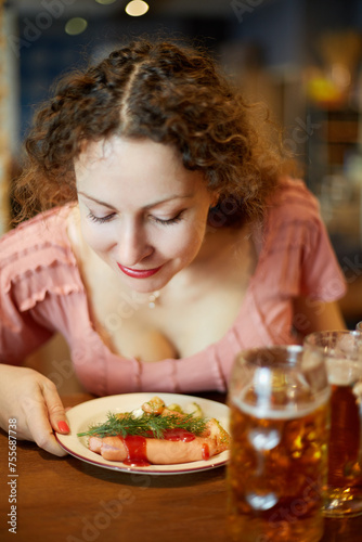 Smiling woman with curly hair sniffs fried sausages with sprig of dill on plate in cafe