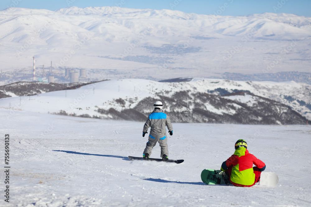 Two snowboarders are on mountain in ski resort, back view, factory far away