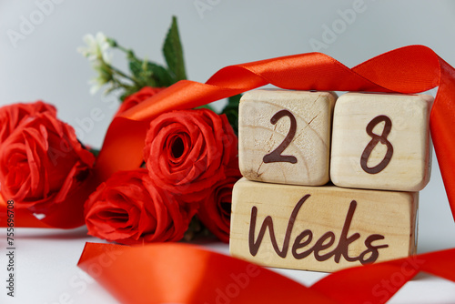 28 week gestational age milestone written on a wooden cube with red roses and ribbons, and a white background