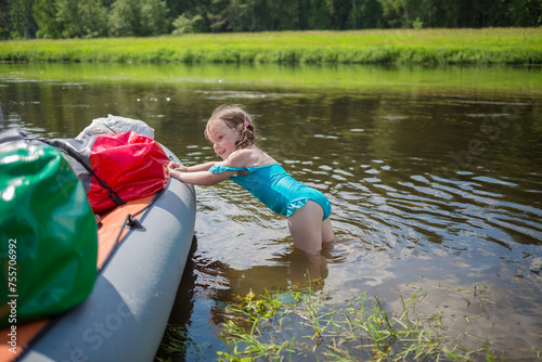 Little girl in swimsuit pushes inflatable boat in river during camping trip at summer photo