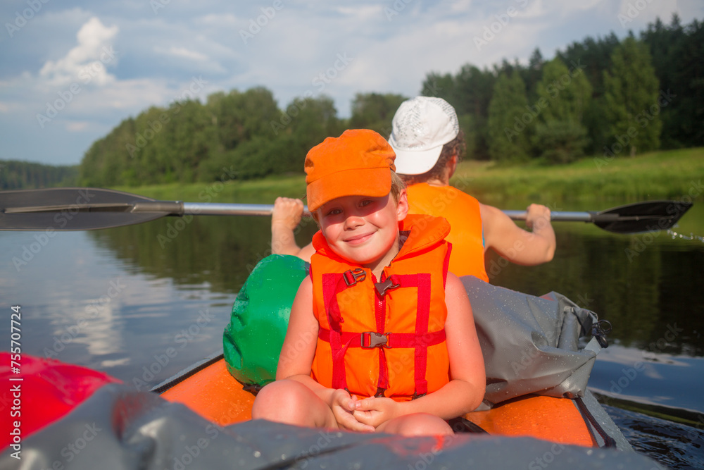 Happy little boy and woman in life-jackets sail on inflatable boat on river at summer, focus on child