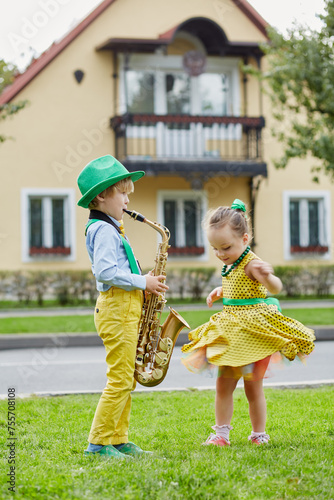 Little boy in dancing suit plays saxophone and little girl dances on grassy lawn against two-storied house photo