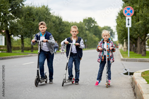 Two boys and girl stand with push scooters on road photo