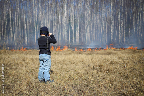 The little boy with the action camera on his head looking at a burning dry grass along the birch grove in the spring photo