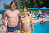 Smiling boy and girl in swimsuit pose near outdoor pool at sunny day, other people out of focus