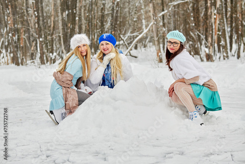 Three smiling women sit squatted near snowdrift at outdoor skate rink in winter park