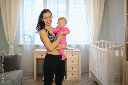 Mother with curly hair in sportswear holding a baby in front of a window in the room