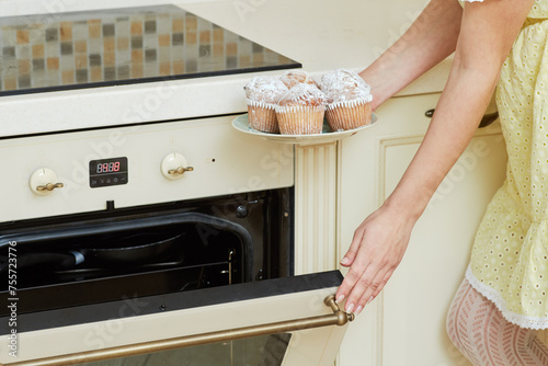 Female hands hold plate with homemade baked cupcakes and close door of oven at kitchen photo
