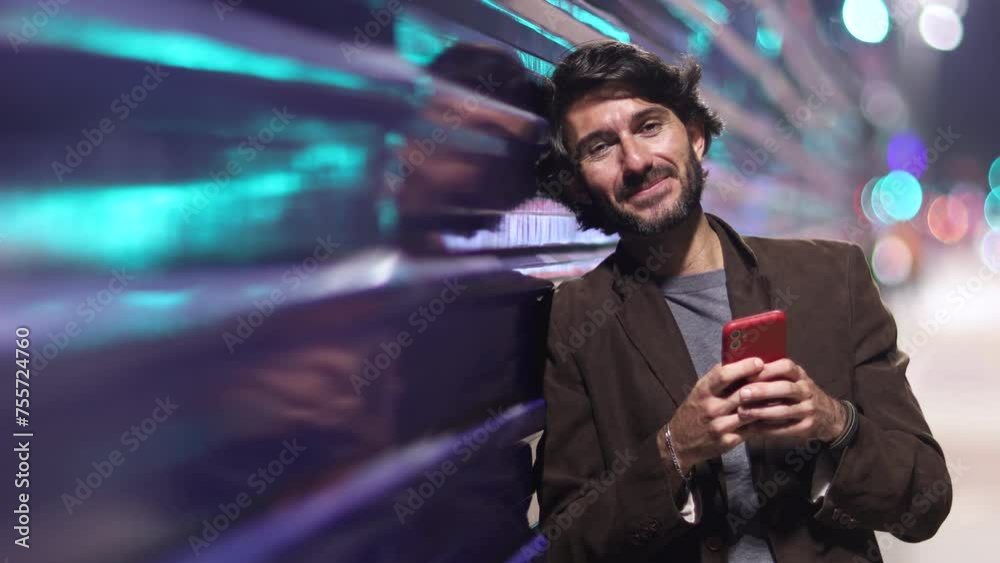 View of young man with smartphone at night time leaning against a blue wall with city view landscape and traffic lights in the background. High quality video. View of young man with smartphone at night time leaning against a blue wall with city view landscape and traffic lights in the background. High quality video.