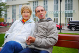 Elderly woman and man sit on bench near building at autumn and look away