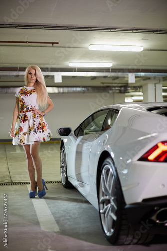 Young blonde woman in flowery dress and blue high-heel shoes stands near modern white car at underground parking