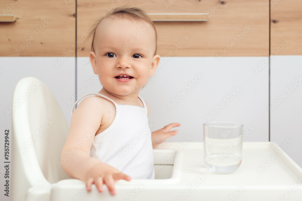 Sweet baby girl holding glass and drinking water. Happy baby sitting in a baby chair in the kitchen. Sweet baby girl holding glass and drinking water. Happy baby sitting in a baby chair in the kitchen.