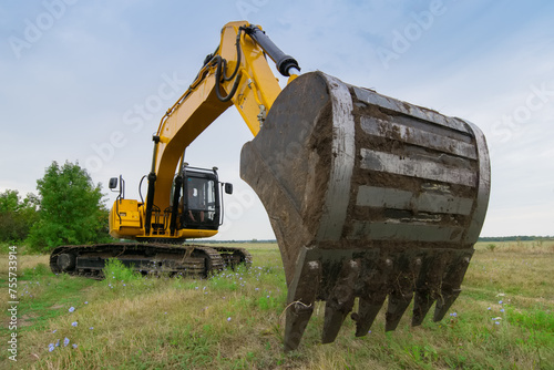 Big yellow excavator in green field at summer day, closeup of excavator bucket