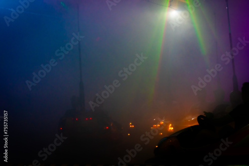 This image depicts the lively and hazy atmosphere of a fairground, focusing on bumper cars amidst a dense fog-like effect. The vibrant beams of light from the overhead ride structure cut through the photo