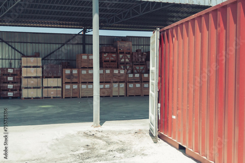 Warehouse area filled with a red container with open doors and wooden crates opposite which processed marble products are placed under a metal roof