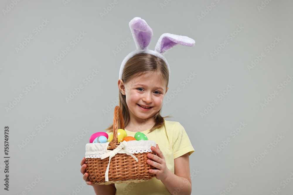 Easter celebration. Cute girl with bunny ears holding basket of painted eggs on gray background