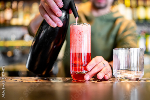 A bartender pours a vibrant red cocktail into a glass at a modern bar, with focus on the drink and hands. Bottles of alcohol line the background, creating an intimate atmosphere.