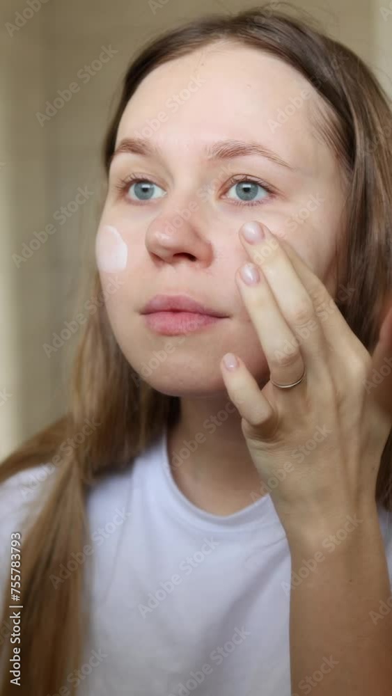 the girl applies the cream to her face and rubs it into her skin. the girl looks in the mirror and applies cream to her skin. vertical video the girl applies the cream to her face and rubs it into her skin. the girl looks in the mirror and applies cream to her skin. vertical video