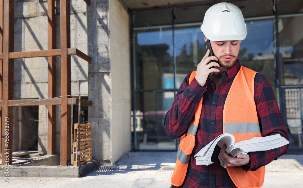 Man builder makes phone call. Guy is employee of architectural bureau. Builder stands near unfinished building. Construction contractor. Man engineer holds stack of documents. Builder calls customer. Man builder makes phone call. Guy is employee of architectural bureau. Builder stands near unfinished building. Construction contractor. Man engineer holds stack of documents. Builder calls customer.