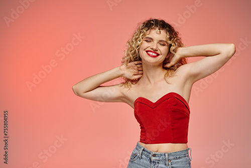 pleased woman with red lips and wavy hair posing in red top with closed eyes on pink and yellow