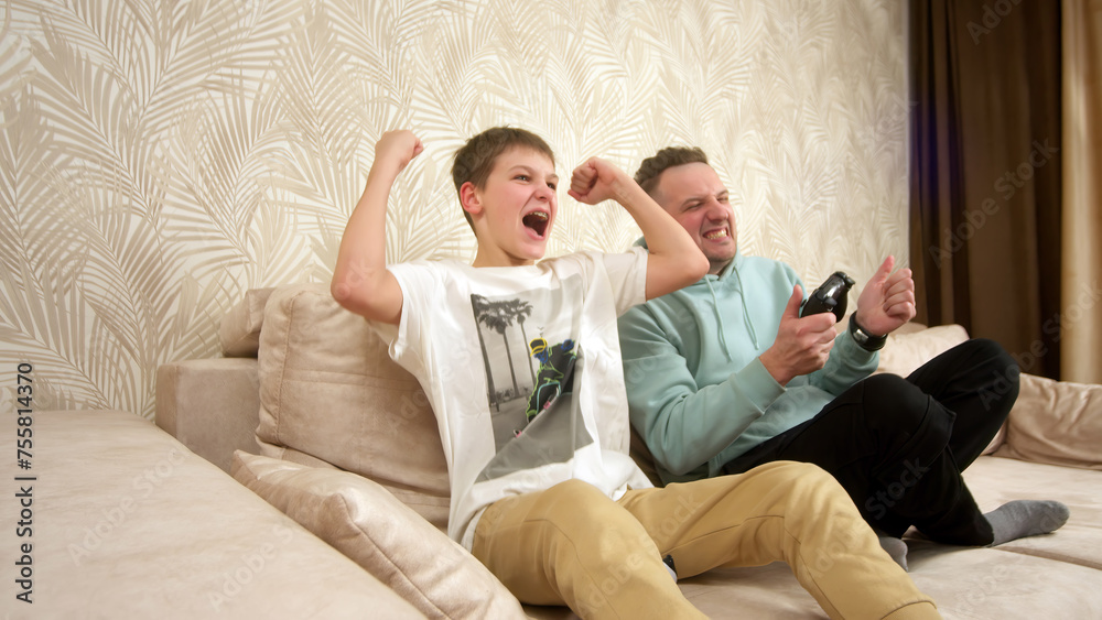 A father spends time with his little son, sitting on the couch and playing a game console. Weekend activities, free time, home entertainment and video games concept. A father spends time with his little son, sitting on the couch and playing a game console. Weekend activities, free time, home entertainment and video games concept.