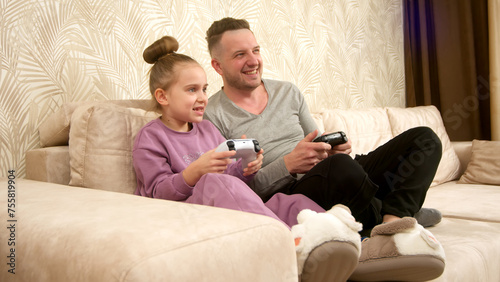 A father spends time with his little daughter, sitting on the sofa and playing a game console. Weekend activities, free time, home entertainment and video games concept. © Vladislav